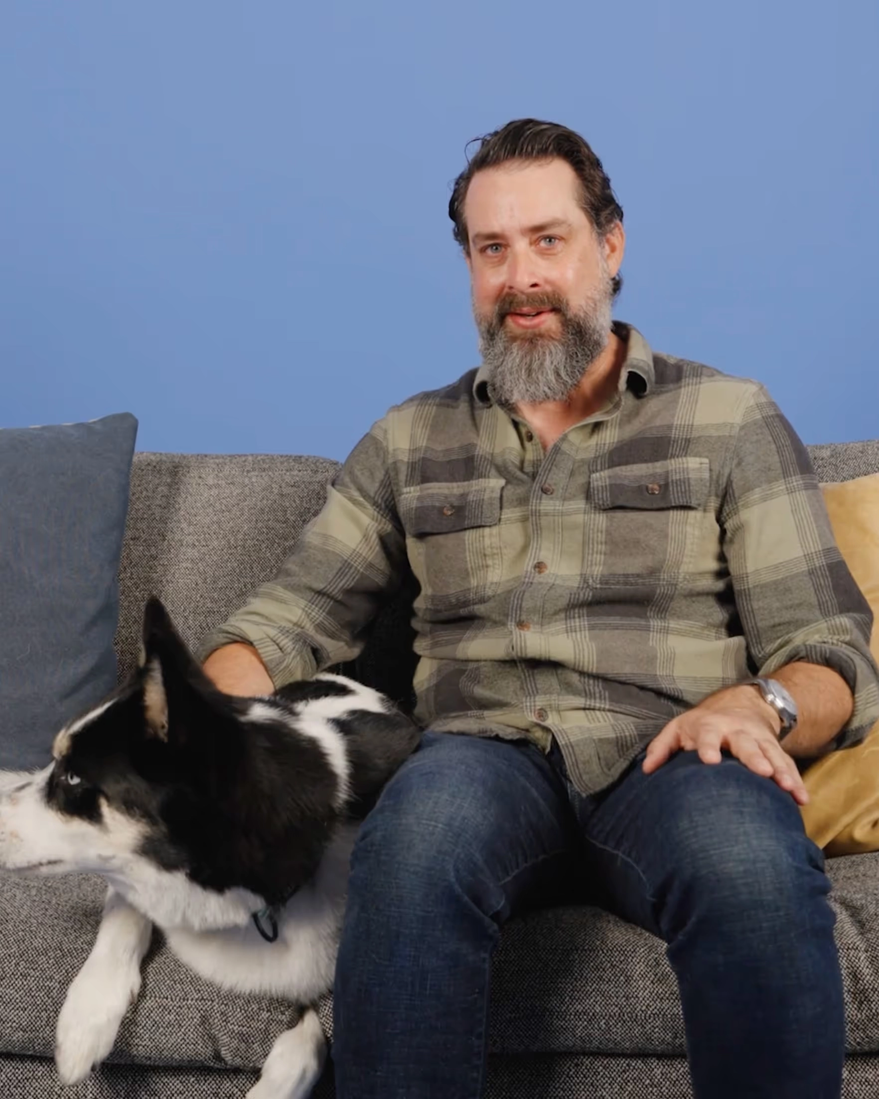 Nick seated on a sofa with a black and white dog, against a blue wall