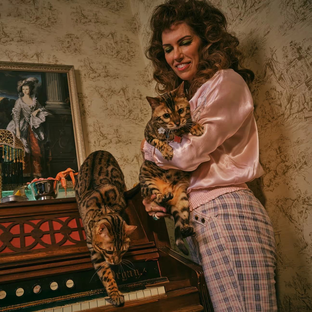 A woman in a pink jacket holding a spotted cat beside an old organ