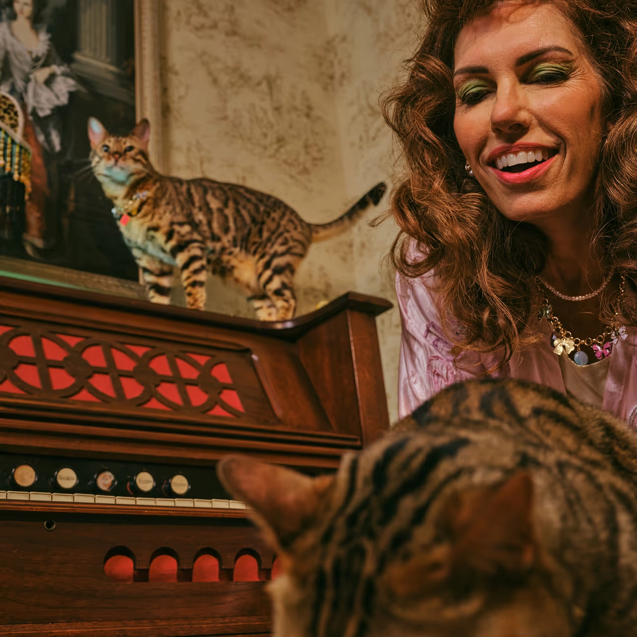 A woman with curly hair smiling over a spotted cat, with a second cat climbing an old organ behind her